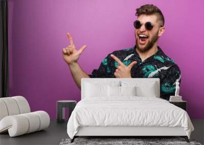 Young man wearing a vacation look pointing with forefingers to a copy space, expressing excitement and desire. Wall mural