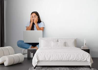 Young mixed race indian woman sitting working on laptop blink through fingers frightened and nervous. Wall mural