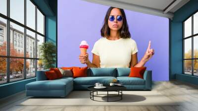 Young mixed race woman eating an ice cream showing number one with finger. Wall mural