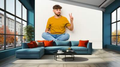 Young Moroccan man sitting on the floor isolated on white background joyful and carefree showing a peace symbol with fingers. Wall mural