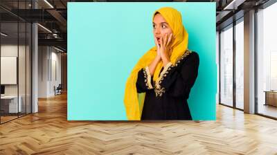 young muslim woman isolated on blue background shouts loud, keeps eyes opened and hands tense. Wall mural