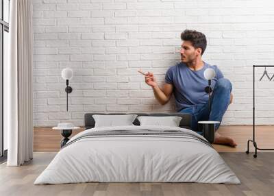 Young natural man sit on a wooden floor pointing to the side, smiling surprised presenting something, natural and casual Wall mural