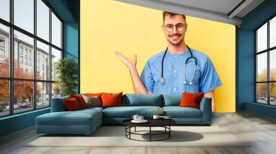 Young nurse caucasian man isolated on yellow background showing a copy space on a palm and holding another hand on waist. Wall mural