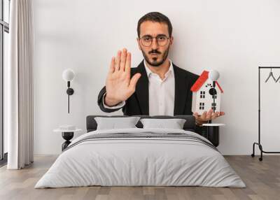 Young real estate agent man holding a model house isolated on white background standing with outstretched hand showing stop sign, preventing you. Wall mural