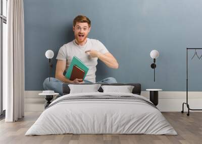 Young redhead student man sitting on the floor surprised, feels successful and prosperous. He is holding books. Wall mural