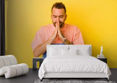 Young tattooed caucasian man isolated on yellow background holding hands in pray near mouth, feels confident. Wall mural