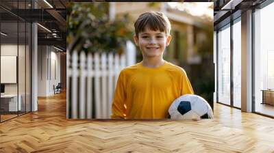 Young boy in yellow shirt smiling and holding a soccer ball, standing in front of a white picket fence on a sunny day, embodying youthful joy and outdoor play Wall mural