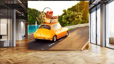 Cute little retro car with suitcases and bicycle on top goes by the road along beautiful harbor between mountain in summer day. Wall mural