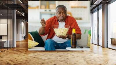 African pensioner sitting at his cozy home, watching television and eating popcorn. He is enjoying the afternoon. Wall mural