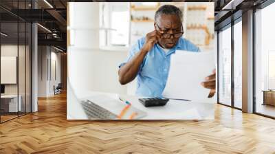 Amazed African pensioner sitting at home and looking at bills he has to pay. He is paying it online over a laptop. Wall mural