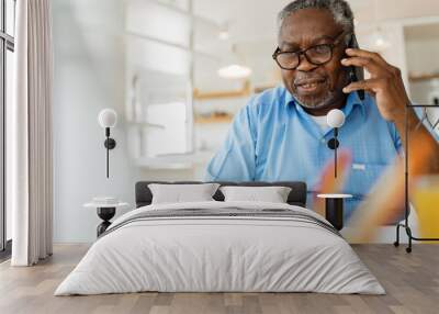 An old black man is sitting at his home office, talking on the phone with a customer support worker and using his laptop. Wall mural