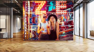 Happy female friends in amusement park drinking beer. Two young women enjoying night at amusement park.  Wall mural