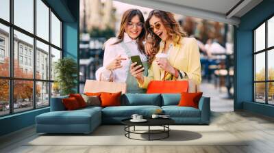 Two happy young women with shopping bags using mobile phone at the street Wall mural