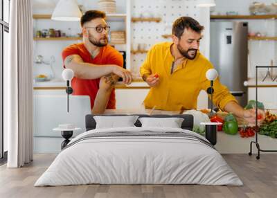 Young gay hipster couple cooking dinner together. A man in the red shirt looking and pointing at the laptop and following the recipe while his partner stirring dinner on the stove. Wall mural