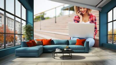 Young woman sitting at the stairs, working on laptop and talking on mobile phone Wall mural