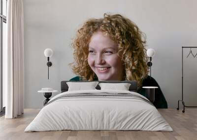 Smiling girl.Young adult girl with curly blonde hair is smiling warmly, wearing a cozy green sweater against a neutral background. The soft lighting enhances teenager's cheerful expression.  Wall mural