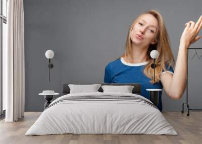 Funny young blonde female shoots in temple, dressed in casual blue t shirt, isolated on gray background with blank space Wall mural