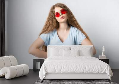 Portrait of charismatic modern girl with curly long hair in casual light clothes and wears round red spectacles posing isolated on white wall background. People lifestyle concept Wall mural