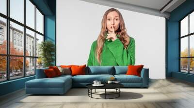 Serious blonde woman shushing, keep quiet gesture, dressed in green cozy sweater, stand over white background Wall mural