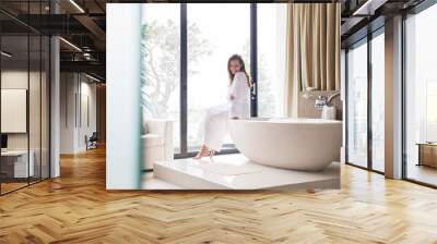 Portrait smiling woman wearing white bathrobe, sitting on edge of bathtub in bathroom Wall mural