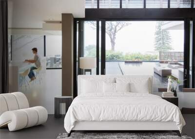 Young man using laptop in modern kitchen, dining area patio door swimming pool in foreground Wall mural