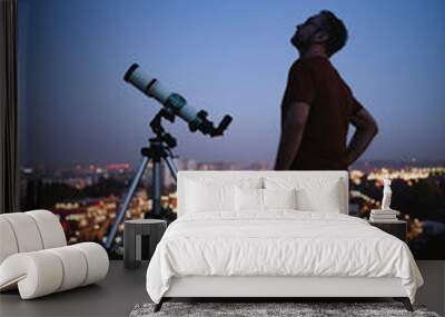 Astronomer with a telescope watching at the stars and Moon with blurred city lights in the background. Wall mural