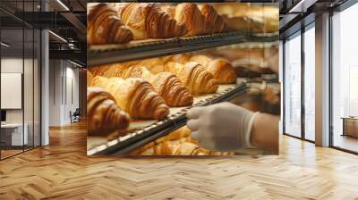 A baker is reaching for a croissant in a bakery. The croissants are displayed on a rack, and the baker is wearing gloves Wall mural