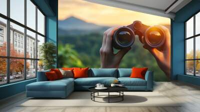 A hand holding binoculars focused on a scenic view. The photo represents the desire to explore and discover new things. Wall mural