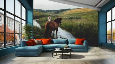 Woman on horseback crossing a stream in a green meadow, with a blurred background of rolling hills Wall mural