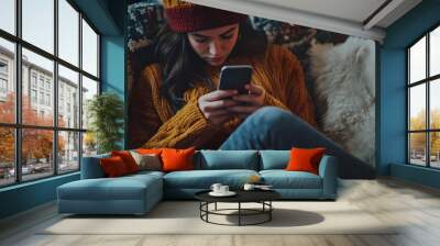 A young woman in a cozy knitted sweater and hat sits on a plush couch, engrossed in her smartphone.  Wall mural