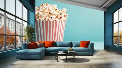 A striped bucket filled with fluffy popcorn, set against a soft teal background, perfect for movie nights or snack cravings. Wall mural