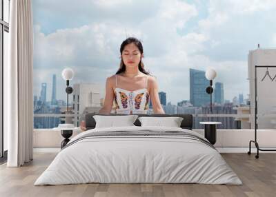 Beautiful Chinese woman wearing sport clothes practices yoga and meditates on top of building with Shanghai skyline background, young female doing yoga asana in city in sunny day. Wall mural