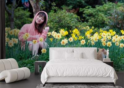 Beautiful young Chinese woman sitting in yellow daffodil flower field smiling with homeless ginger tabby cat. Candid happy moment. Emotions, people, beauty, youth and lifestyle portrait. Wall mural