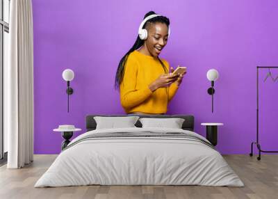 African American woman wearing headphones listening to music from smart phone on colorful purple isolated studio background Wall mural