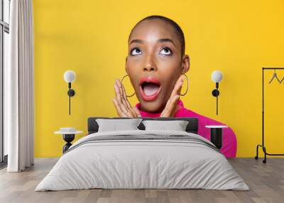 Close up portrait of surprised young African American woman looking upward with hands cupped around mouth isolated on studio yellow background Wall mural