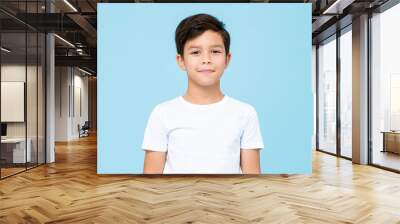 Cute smiling boy in plain white t shirt looking at camera in isolated studio light blue color background Wall mural