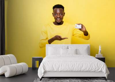 excited african man in casual attire holding and presenting credit card on yellow studio background Wall mural