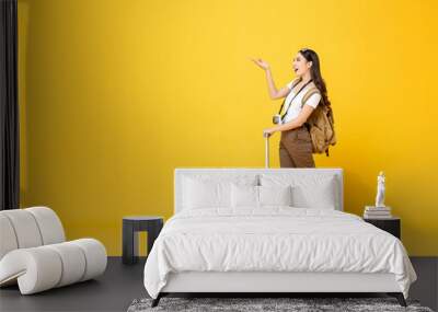 Full length portrait of beautiful smiling young Asian woman tourist with trolley bag looking and opening hand to copy space isolated on colorful yellow studio background Wall mural