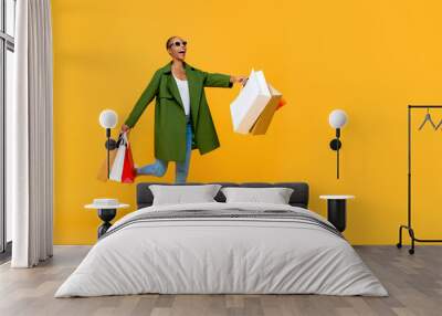 Full length portrait of cheerful attractive African American woman holding shopping bags while prancing in isolated studio yellow background Wall mural