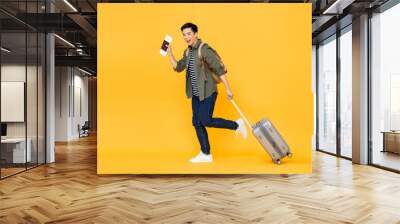 Full length portrait of cheerful young smiling tourist Asian man walking while holding luggage and showing passport with ticket in isolated studio yellow background Wall mural