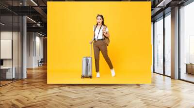 Full length portrait of smiling cute young Asian woman tourist with luggage isolated on yellow studio background Wall mural