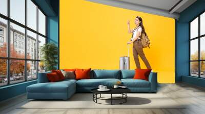 Full length travel concept portrait of beautiful smiling young Asian woman tourist with trolley bag pointing up isolated on colorful yellow studio background Wall mural