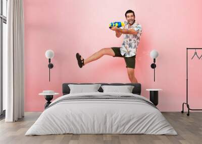 Handsome smiling young Caucasian tourist man playing with water gun and jumping in studio pink background for Songkran festival in Thailand and southeast Asia Wall mural