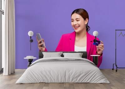 Happy beautiful young Asian woman in pink blazer shopping with credit card via mobile phone isolated on purple background Wall mural