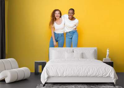 Happy two interracial woman friends smiling and holding each other in isolated studio yellow color background Wall mural