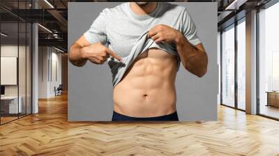 Muscular male model lifting up t-shirt to show abs while standing on gray isolated background in studio Wall mural