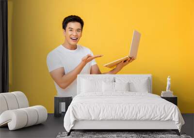 Portrait of smiling young handsome Asian man presenting laptop while smiling and looking  at camera in isolated studio yellow background Wall mural