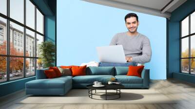 Portrait of smiling young handsome Caucasian man looking at camera while sitting using laptop in isolated studio blue background Wall mural