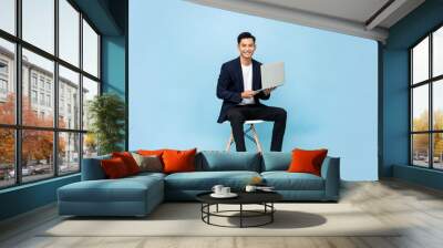 Portrait of smiling young handsome southeast Asian millenial businessman with laptop sitting on chair in light blue studio background Wall mural