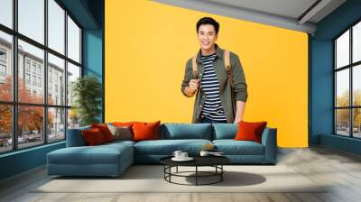 Portrait of smiling young male Asian tourist carrying backpack ready to travel in isolated studio yellow background Wall mural